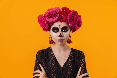 Cadılar Bayramı makyajı ve çiçek çelengi giymiş güzel bir kız sarı duvarın üzerinde izole edilmiş kameraya poz veriyor.