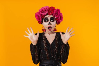 Cadılar Bayramı makyajı ve çiçek çelengi giymiş şok olmuş bir kız sarı duvarın üzerinden izole edilmiş kameraya poz veriyor.