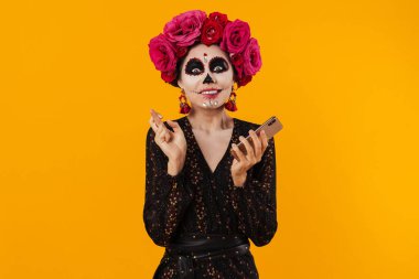 Cadılar Bayramı makyajlı heyecanlı kız sarı duvarda izole edilmiş cep telefonu kullanırken parmaklarını bir arada tutuyor.