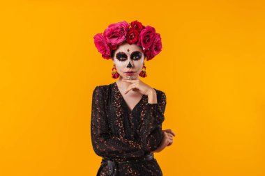 Cadılar Bayramı makyajı ve çiçek çelengi giymiş güzel bir kız sarı duvarın üzerinde izole edilmiş kameraya poz veriyor.