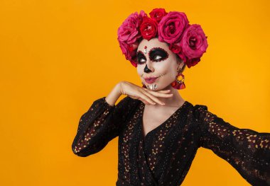 Cadılar bayramı makyajı ve çiçek çelengi giymiş mutlu bir kız sarı duvarda izole edilmiş selfie fotoğrafı çekiyor.