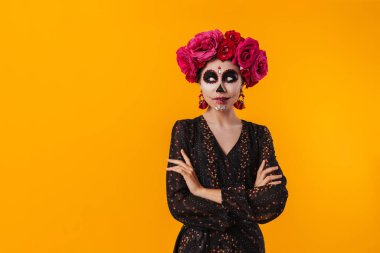 Cadılar Bayramı makyajlı ve çiçek çelenkli kız sarı duvarda izole edilmiş kameraya poz veriyor.