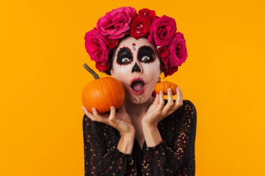 Cadılar Bayramı makyajlı genç kız sarı arka planda izole edilmiş balkabaklarıyla poz veriyor.