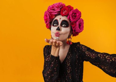 Cadılar Bayramı makyajlı mutlu bir kız, sarı duvarın üzerinden selfie çekerken havada öpüşüyor.