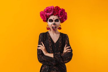 Cadılar Bayramı makyajı ve çiçek çelengi giymiş ürkütücü kız sarı duvarda izole edilmiş kameraya bağırıyor.