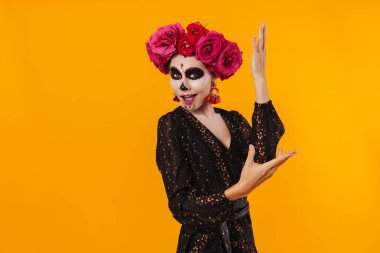 Cadılar bayramı makyajı ve çiçek çelengi giymiş neşeli kız sarı duvarın üstünde dans ediyor.