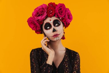 Cadılar bayramı makyajı ve çiçek çelengi giymiş güzel bir kız sarı duvarın üstünde cep telefonuyla konuşuyor.