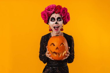 Cadılar Bayramı makyajlı heyecanlı genç kız sarı arka planda izole edilmiş balkabağıyla poz veriyor.