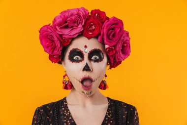 Cadılar Bayramı makyajı ve çiçek çelengi giymiş şok olmuş bir kız sarı duvarın üzerinden izole edilmiş kameraya poz veriyor.