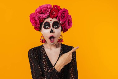 Cadılar Bayramı makyajı ve çiçek çelengi giymiş şaşkın bir kız sarı duvarın üzerinde izole edilmiş bir şekilde parmağını işaret ediyor.