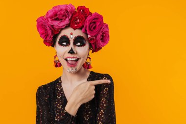 Cadılar Bayramı makyajlı heyecanlı kız ve çiçek çelengi sarı duvarın üzerinde izole edilmiş parmakları işaret ediyor.