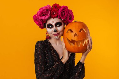 Cadılar Bayramı makyajlı mutlu genç kız sarı arka planda izole edilmiş balkabağıyla poz veriyor.