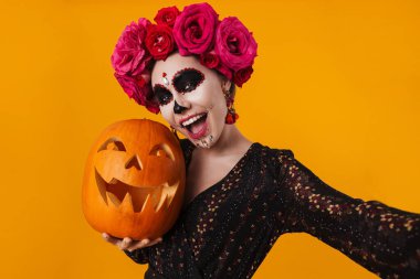 Cadılar Bayramı makyajlı neşeli kız sarı arka planda balkabağıyla poz verirken selfie çekiyor.