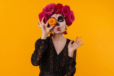 Cadılar Bayramı makyajlı neşeli genç kız sarı arka planda izole edilmiş küçük balkabağıyla dalga geçiyor.