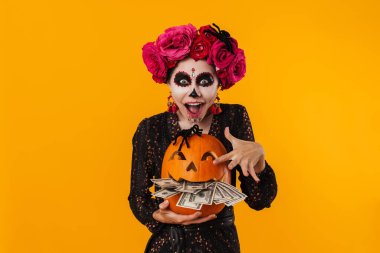 Cadılar Bayramı makyajlı keyifli beyaz kız sarı duvarın üzerinde balkabağı ve dolarlarla poz veriyor.