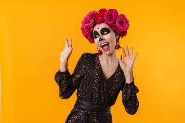 Cadılar bayramı makyajı ve çiçek çelengi giymiş heyecanlı kız sarı duvarda izole edilmiş kameraya poz veriyor.