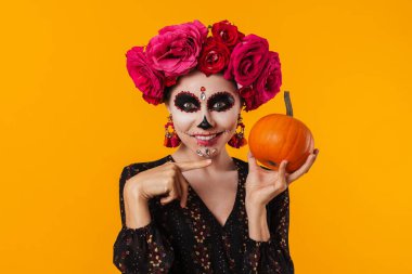 Cadılar Bayramı makyajlı mutlu genç kız sarı arka planda izole edilmiş balkabağını işaret ediyor.