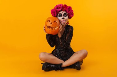 Cadılar Bayramı makyajlı genç bir kız, sarı arka planda izole bir şekilde otururken balkabağı dilini gösteriyor.