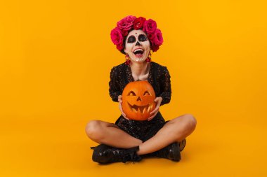 Cadılar Bayramı makyajlı neşeli genç kız, sarı arka planda izole bir şekilde balkabağı yerde oturuyor.