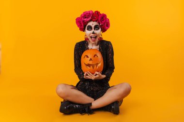 Cadılar Bayramı makyajlı keyifli Kafkasyalı kız. Sarı arka planda, kabağı yerde izole bir şekilde oturuyor.