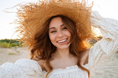 Hasır şapkalı, gülümseyen ve dışarıdaki kameraya bakan beyaz, çekici, esmer kadın.