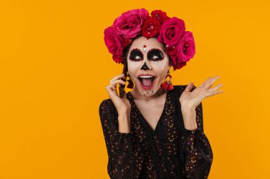 Cadılar bayramı makyajı ve çiçek çelengi giymiş heyecanlı kız sarı duvarda izole bir şekilde cep telefonundan konuşuyor.