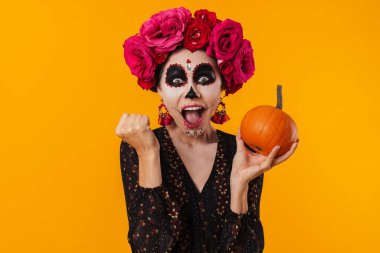 Cadılar Bayramı makyajlı heyecanlı kız sarı arka planda saklanmış balkabağıyla poz verirken kazanan hareketi yapıyor.