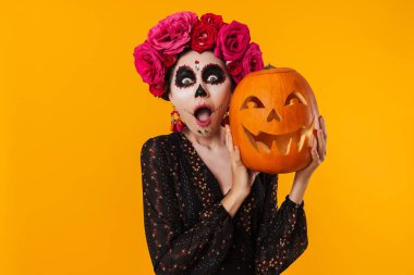 Cadılar Bayramı makyajlı genç kız sarı arka planda izole edilmiş balkabağıyla poz veriyor.