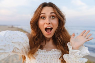 Beyaz tenli, heyecanlı, esmer kadın kameraya bakıp, sahilde selfie çekiyor.