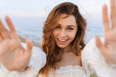 Beyaz tenli çekici esmer kadın gülümsüyor ve kumsalda avuçlarını kameraya gösteriyor.