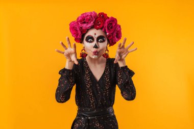 Cadılar bayramı makyajı ve çiçek çelengi giymiş ürkütücü kız sarı duvarda izole edilmiş kameraya poz veriyor.