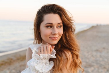 Genç beyaz tenli çekici esmer kadın gülümsüyor ve kumsala bakıyor.