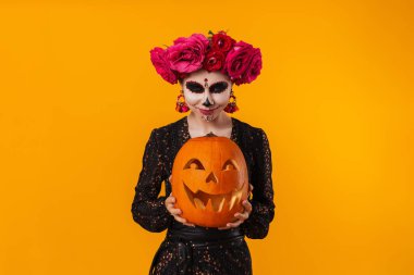 Cadılar Bayramı makyajlı mutlu genç kız sarı arka planda izole edilmiş balkabağıyla poz veriyor.