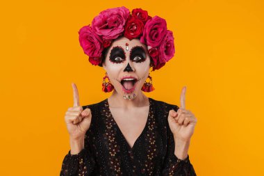 Cadılar bayramı makyajı ve çiçek çelengi giymiş heyecanlı kız sarı duvarın üzerinde izole bir şekilde parmaklarını yukarı kaldırıyor.