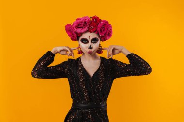 Cadılar Bayramı makyajlı kız ve çiçek çelengi sarı duvarın üzerinde parmaklarını kafasına doğru gösteriyor.