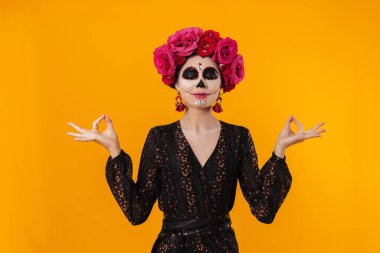 Cadılar Bayramı makyajı ve çiçek çelengi giymiş mutlu bir kız sarı duvarın üzerinde izole edilmiş bir şekilde meditasyon yapıyor.