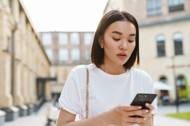 Genç, güzel Asyalı bir kadın sokakta yürürken telefon kullanıyor.