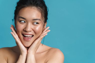 Mavi arka planda tek başına dikilirken gülümseyen üstsüz bir Asyalı kadının güzel portresi.