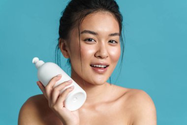 Mavi arka planda soyutlanmış, boş plastik şişeyi gösteren, gülümseyen üstsüz, genç Asyalı bir kadının güzel portresi.