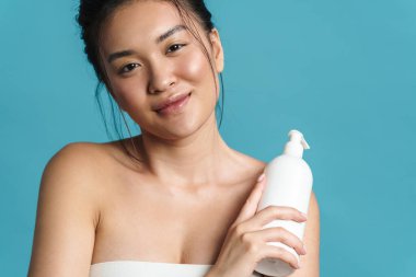Mavi arka planda soyutlanmış, boş plastik şişeyi gösteren, gülümseyen üstsüz, genç Asyalı bir kadının güzel portresi.