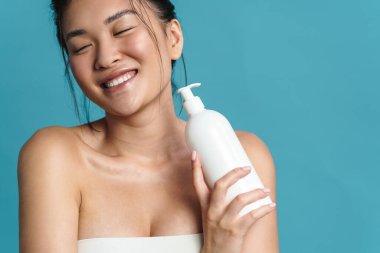 Mavi arka planda soyutlanmış, boş plastik şişeyi gösteren, gülümseyen üstsüz, genç Asyalı bir kadının güzel portresi.