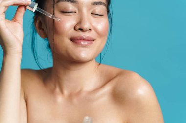 Mavi arka planda izole edilmiş yüz serumunu uygulayan mutlu üstsüz Asyalı kız görüntüsü