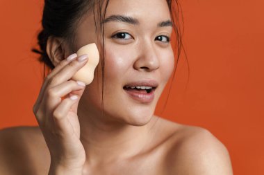 Turuncu arka planda izole edilmiş kozmetik sünger kullanan güzel üstsüz Asyalı kız görüntüsü