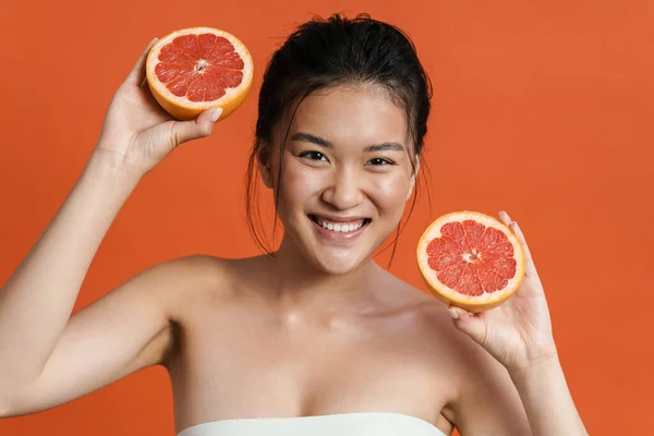オレンジ色の背景に隔離されたまま立ちながらポーズをとる陽気な若いアジアの女性の美しさの肖像画 カットグレープフルーツを保持 — ストック写真
