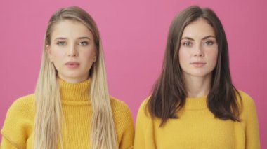 İki şaşırmış kadının yakın çekim görüntüsü stüdyodaki pembe arka planda tek başına duran kameraya bakıyor.
