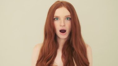 Şok olmuş bir kadın kameraya bakıyor. Stüdyoda bej arka planda tek başına duruyor.
