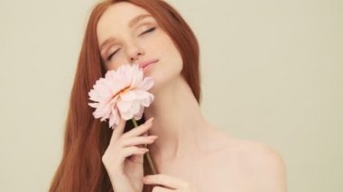 Muhteşem bir kadın elinde çiçekle kameraya poz veriyor. Stüdyoda bej arka planda tek başına duruyor.