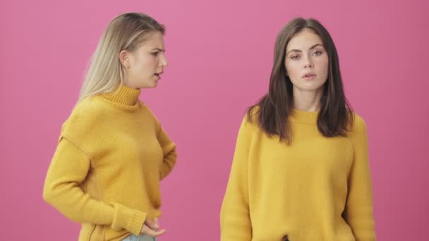 黄色のセーターを身に着けているブロンド女性ありますScading別のブルネット女性立って絶縁上のピンクの背景でスタジオ — ストック動画