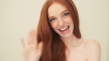 Pozitif bir kadın elini kameraya doğru sallıyor. Stüdyoda bej arka planda tek başına duruyor.