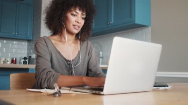 Mutlu bir Afrikalı kadın evde oturmuş kulaklıkla müzik dinlerken dizüstü bilgisayar kullanıyor.
